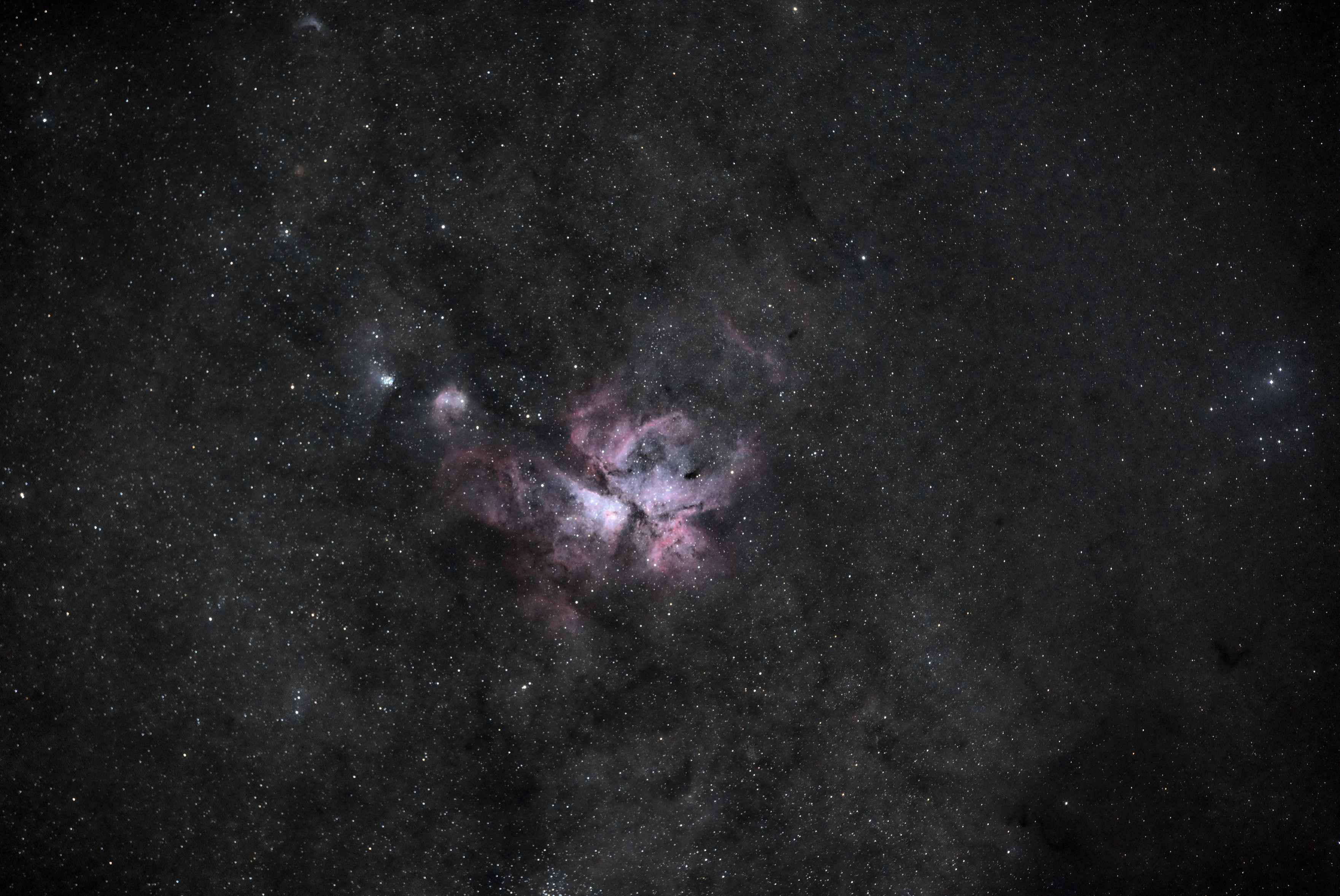 A purple-ish pink-ish blob of nebula, with dark lanes crossing over it. Smaller tufts of nebulously sit up and to the left. On the right hand side a cluster of stars is surrounded by a faint blue glow.

Wisps of dark dust cross the entire faintly glowing background.