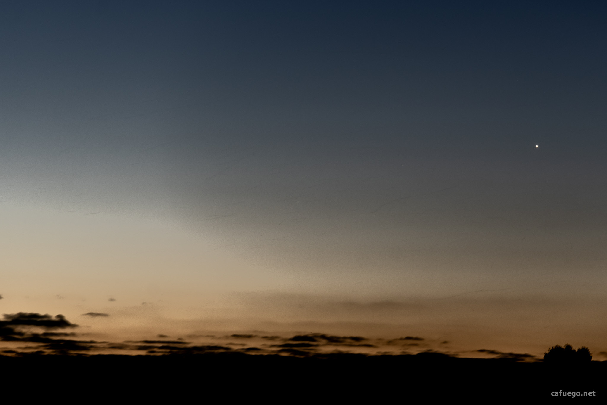 A sunset gradient sky, orange at bottom and blue at top with a small fuzzy blob in the middle (the comet). At top right, Jupiter is a bright circle flanked by two small moons. Weird diagional dark streaks run across the whole image - flying fox trails.