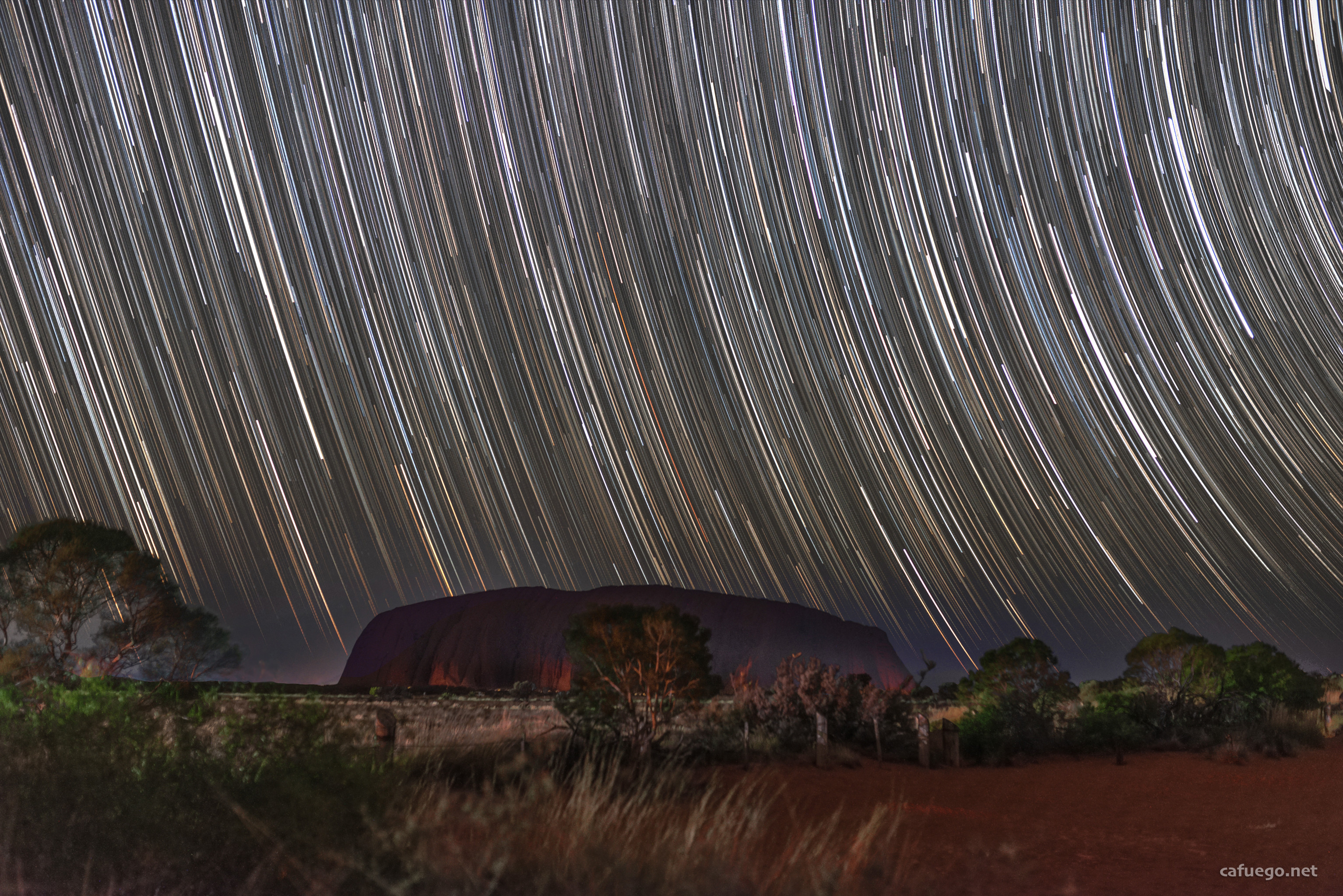 Uluru (Ayers Rock) at night with some red dust and shrubs in the foreground. Beyond it, the spinning earth has turned the dots of stars into long arcs.